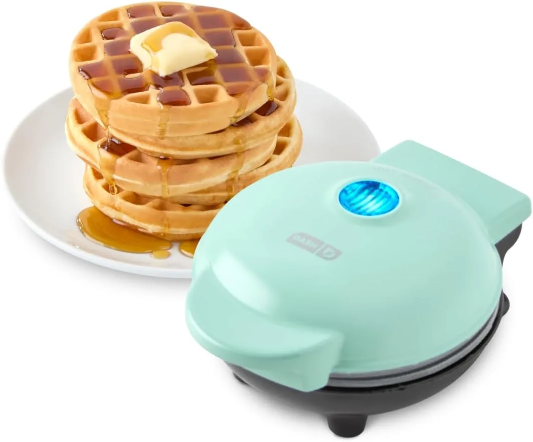 Small kitchen gadgets that look expensive arranged on a sleek countertop: chopper, gold scissors, bamboo board, aqua waffle maker and clear hobnail glasses